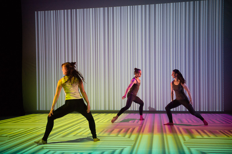 Dancers assume wide positions with bent legs. A pattern of white lines is projected on the back wall and floor